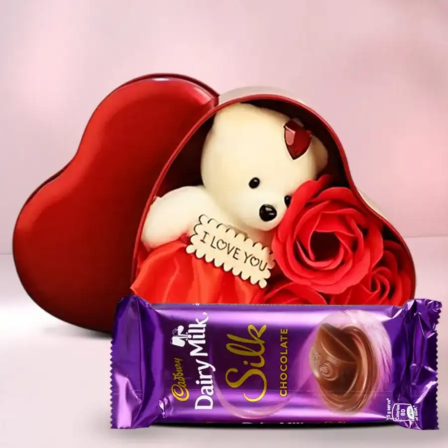 Heart-shaped box filled with fresh roses, a cuddly teddy bear, and a Cadbury Silk chocolate bar, arranged for a romantic gift.