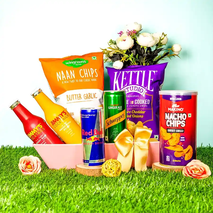 A beautifully arranged hamper featuring various drinks including Schweppes Ginger Ale, Jimmys Mango Chilli Mojito, Whisky Sour mix, and Red Bull alongside snacks like Wingreens Butter Garlic Naan Chips, Kettle Studio Cheddar & Red Onion Chips, and Makino Sweet Chilli Nachos, perfect for cocktails and munching.