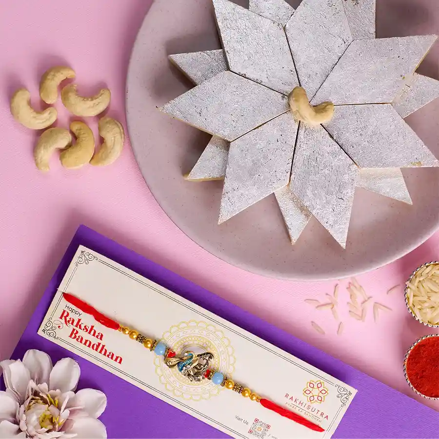 Designer Kundan Rakhi accompanied by a box of Kaju Katli, Roli, Tika, Chawal, and a heartfelt message card, arranged on a decorative surface for Raksha Bandhan.